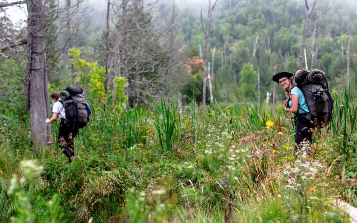 two outward bound students carrying backpacks hike through a green landscape